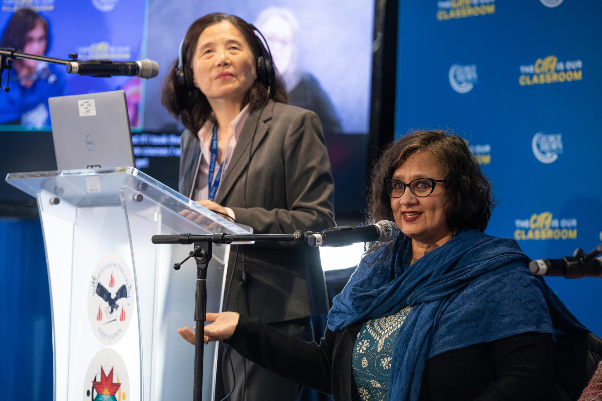 A woman at a podium with another seated next to her with a microphone