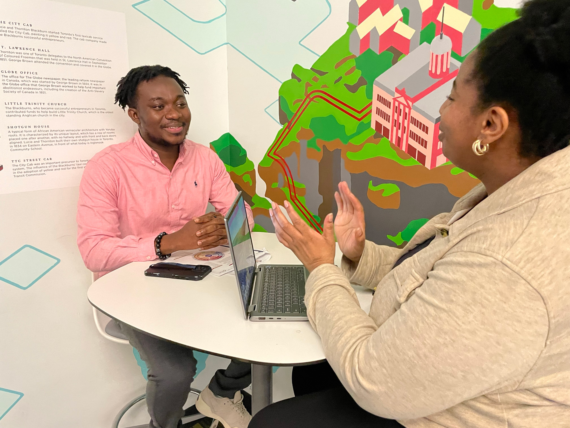 Two people sit at a table chatting with an open laptop and colourful background