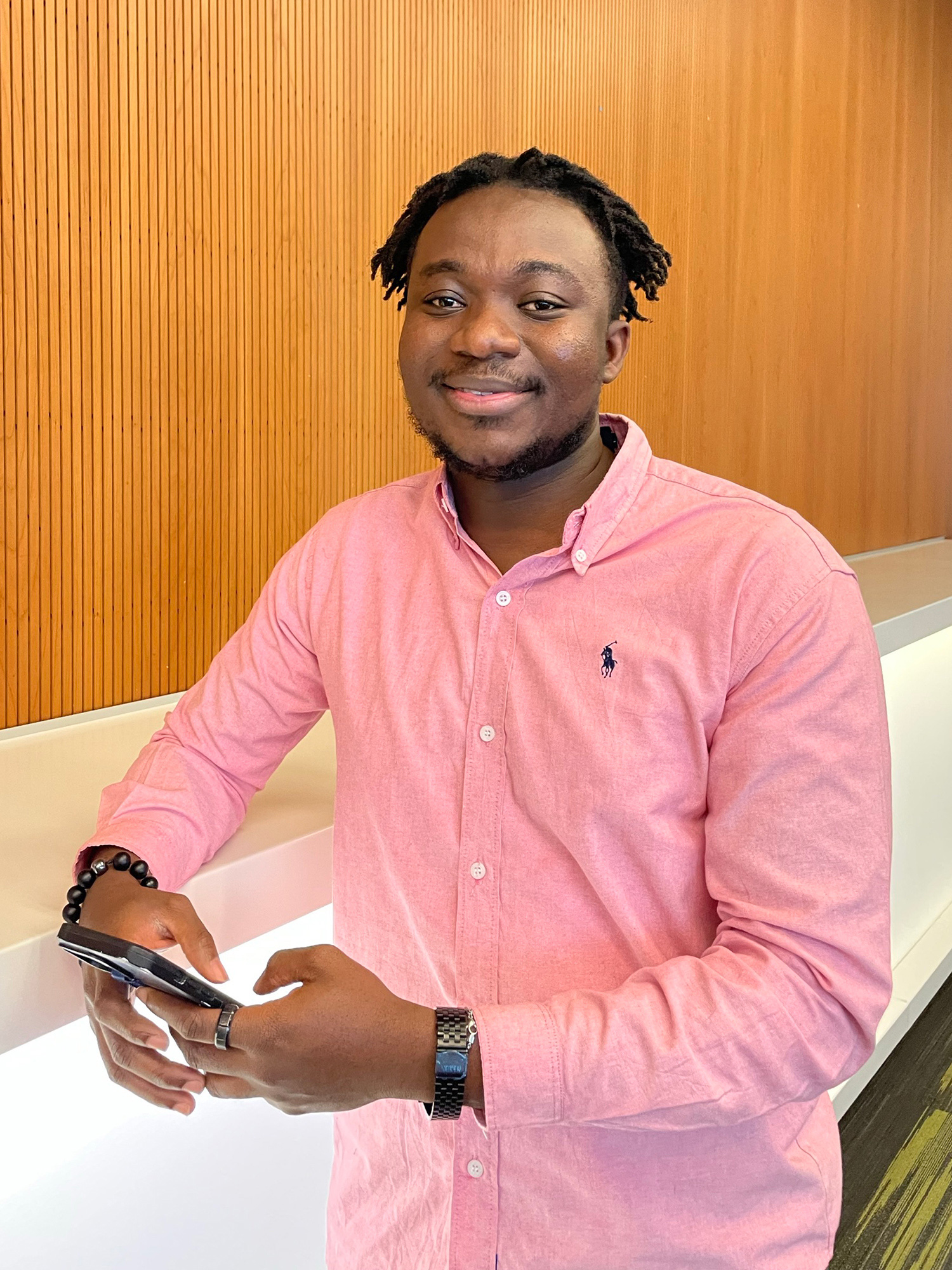 A Black man in a pink shirt holds a mobile device and smiles at the camera