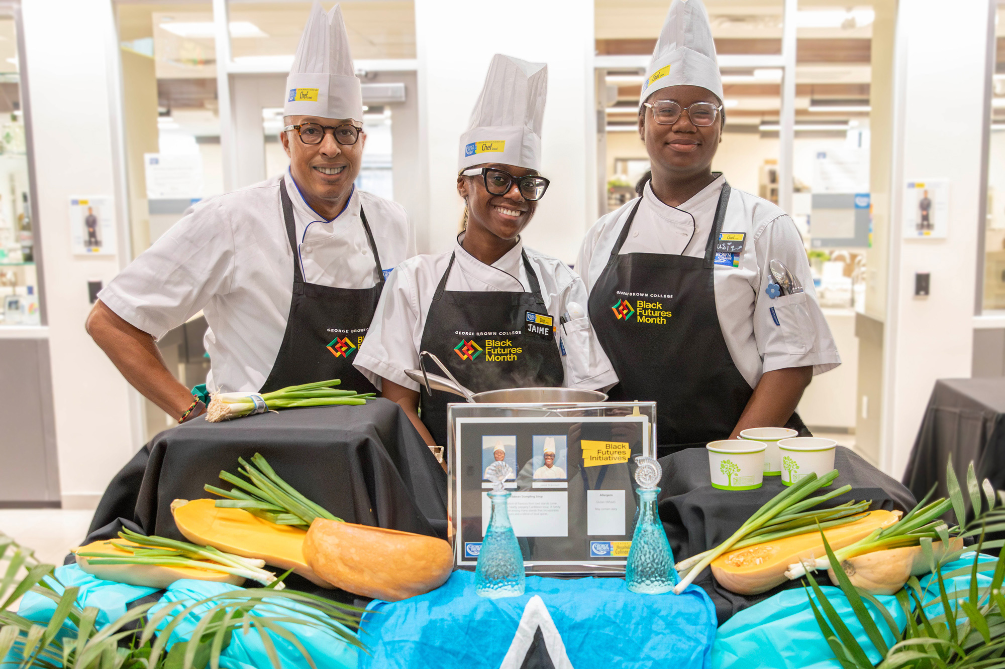 Three chefs in chef's uniforms stand behind a food station and smile