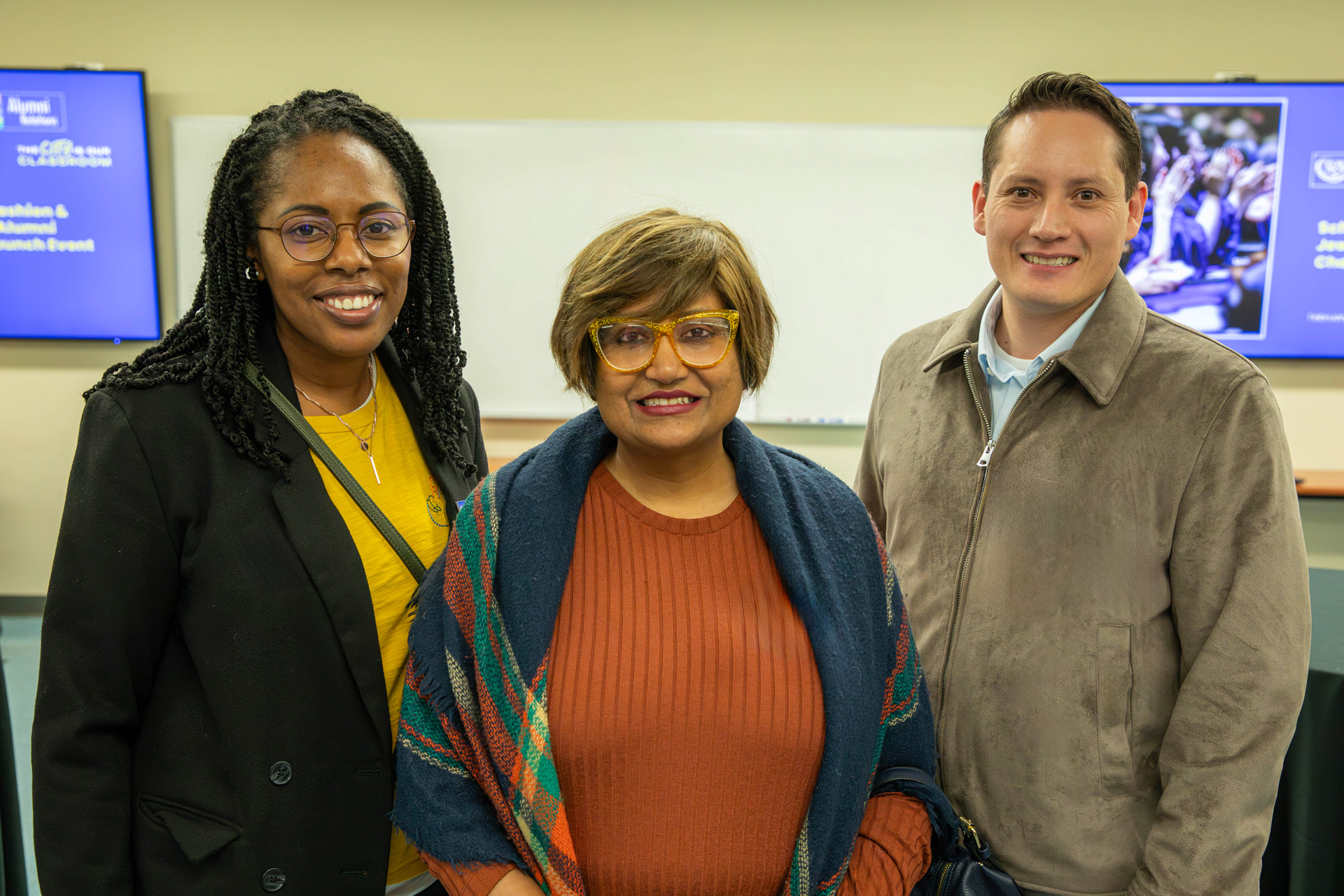 Three individuals standing in front of digital screens with alumni chapter information.