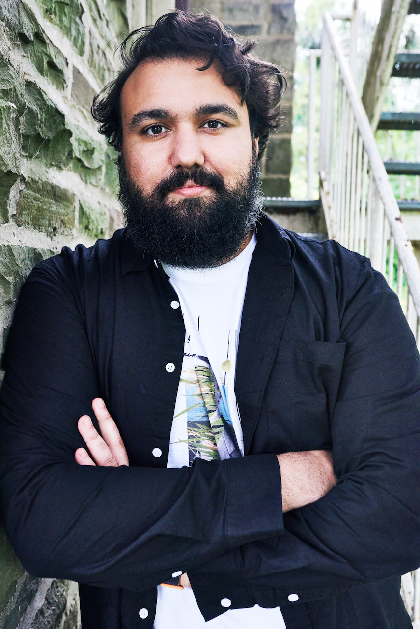 Man in a black shirt leans against a stone wall with arms crossed